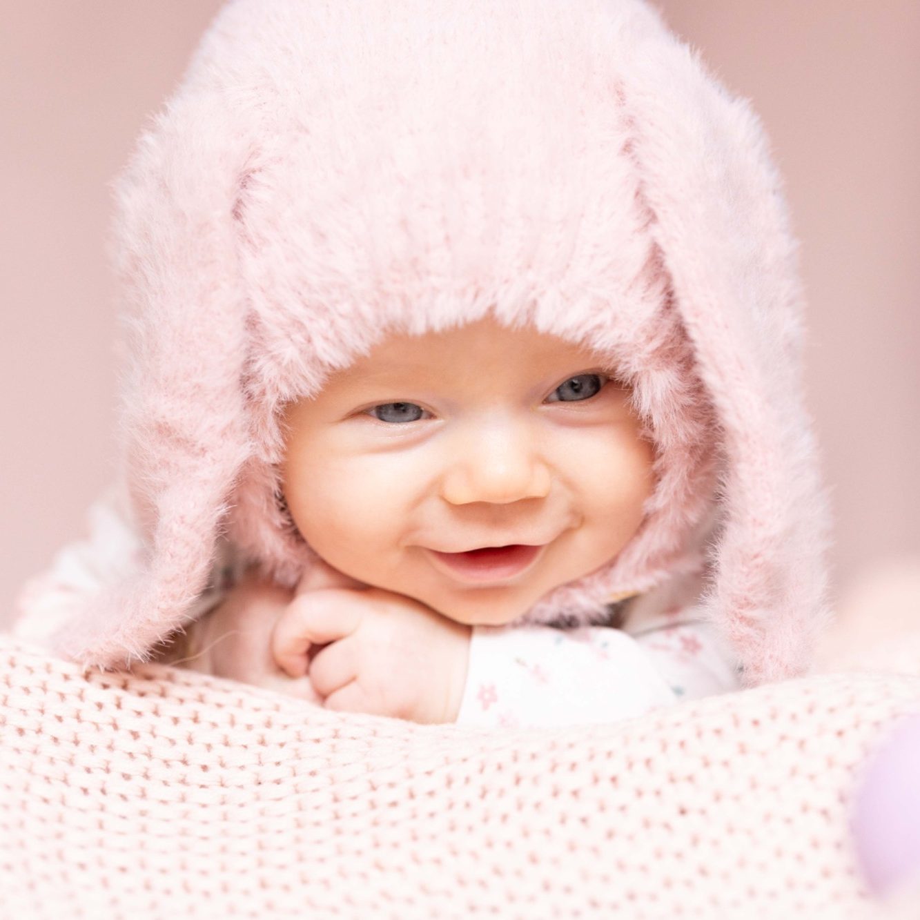 Ein lächelndes Baby trägt eine rosa Mütze mit Ohren und schaut fröhlich in die Kamera.
